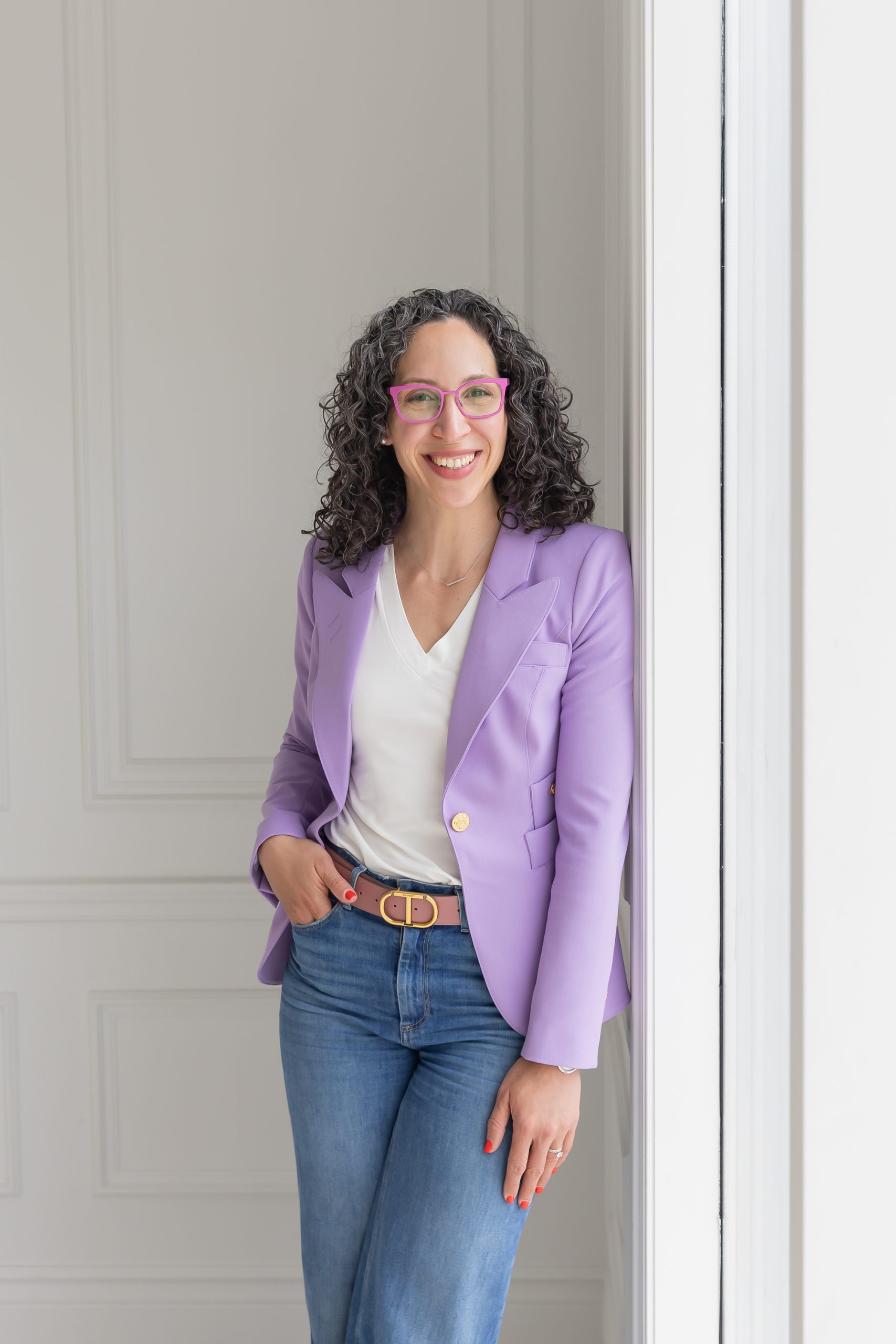 Smiling woman with curly hair wearing a burgundy blazer and white blouse against a dark gray wall.