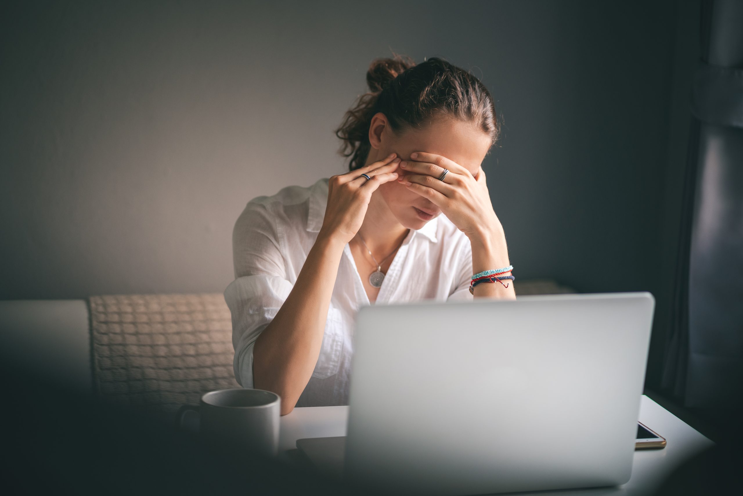 Young woman in a white shirt sits at a table in front of a laptop, her eyes and head ache, workaholism, stress burnout.