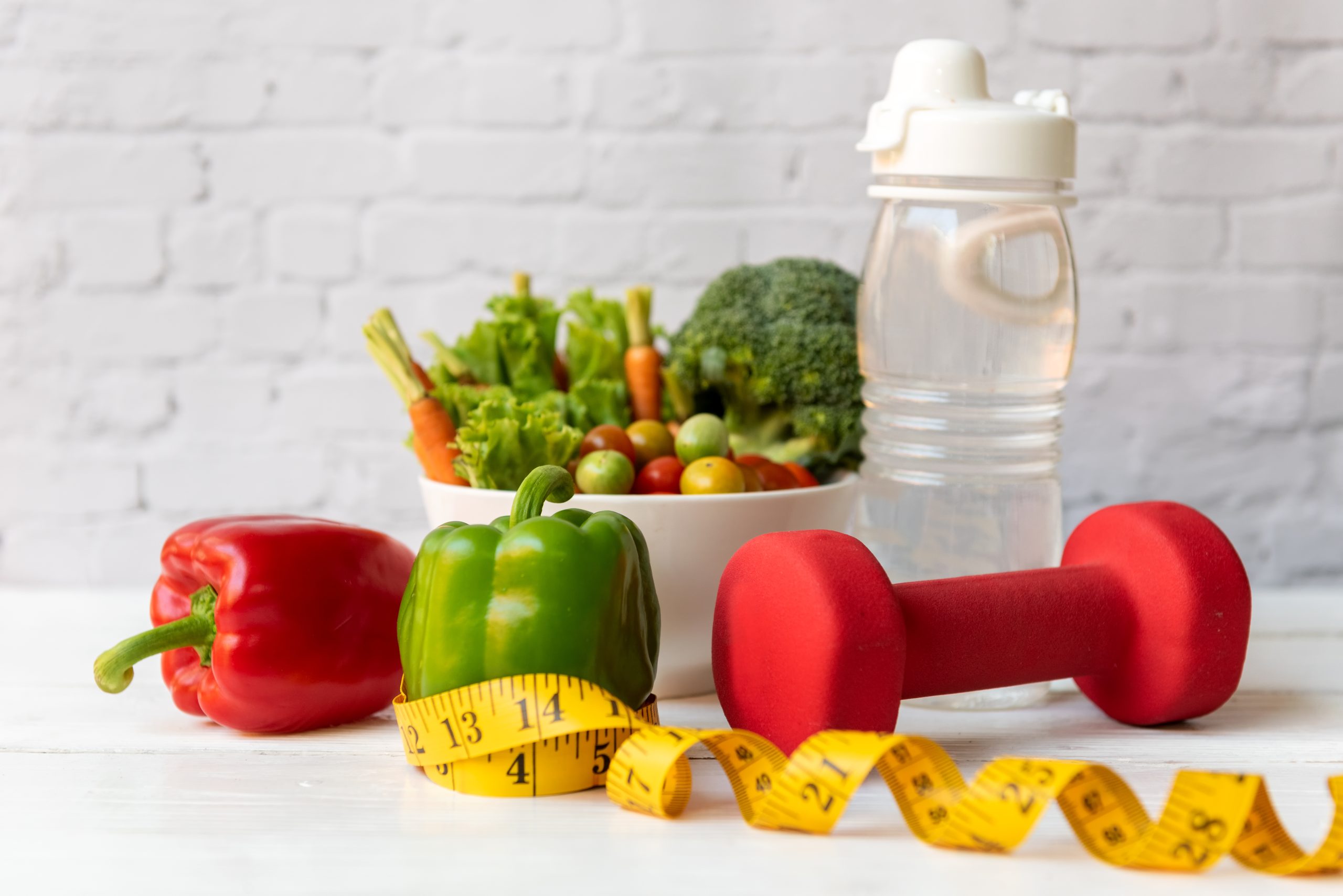 Fresh vegetables, water bottle, red dumbbell, and measuring tape on a white table.