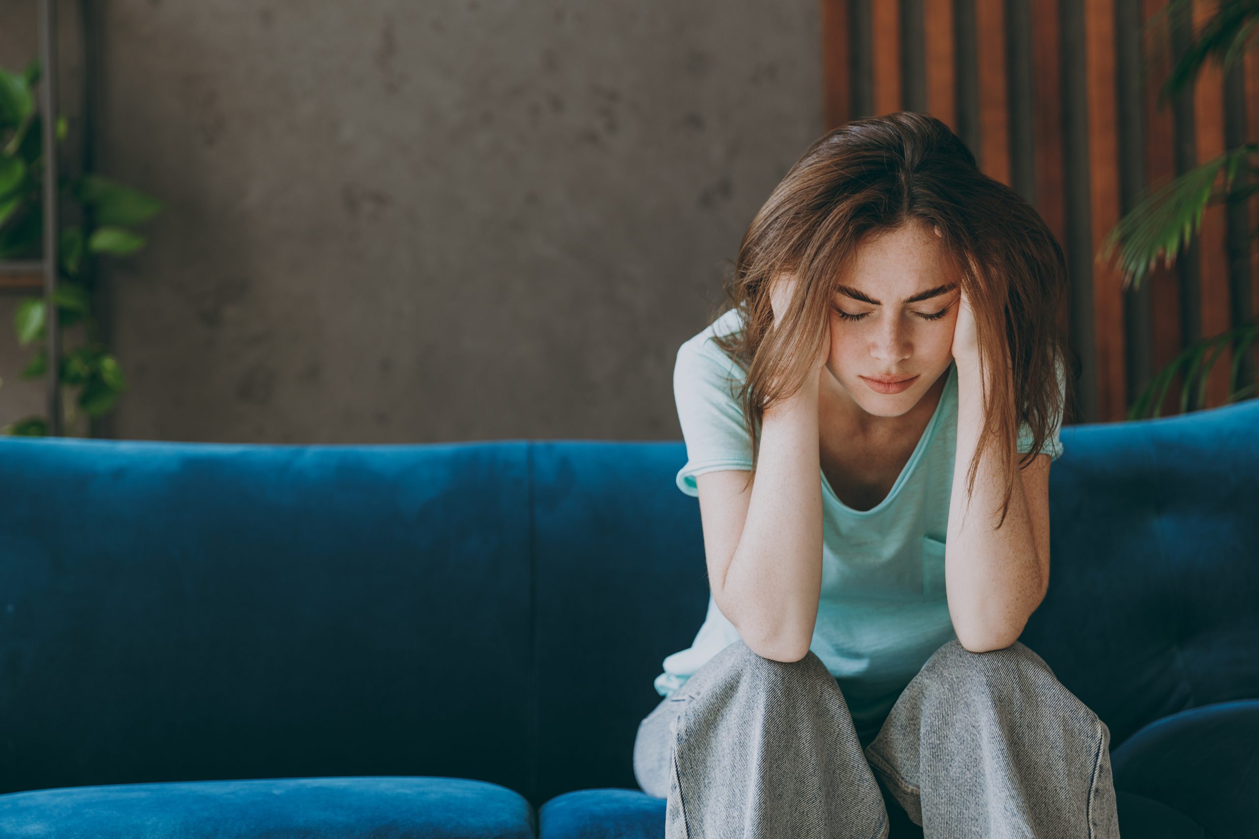 Young frustrated ill woman she 20s wearing casual clothes mint t-shirt holding head suffer from headache close eyes sit on blue sofa indoor rest at home in own room apartment