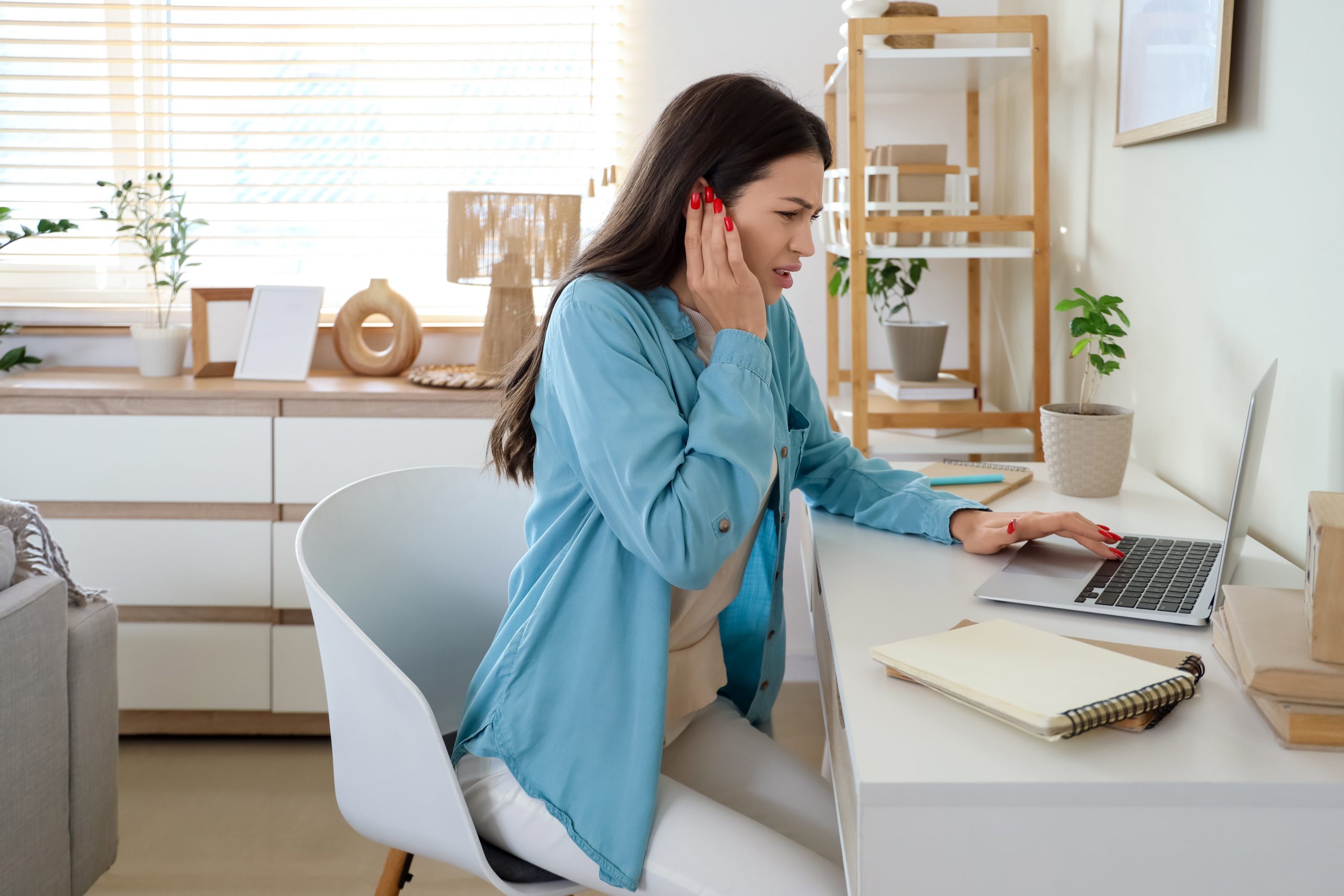 Woman holding her painful ear while using a laptop at home.