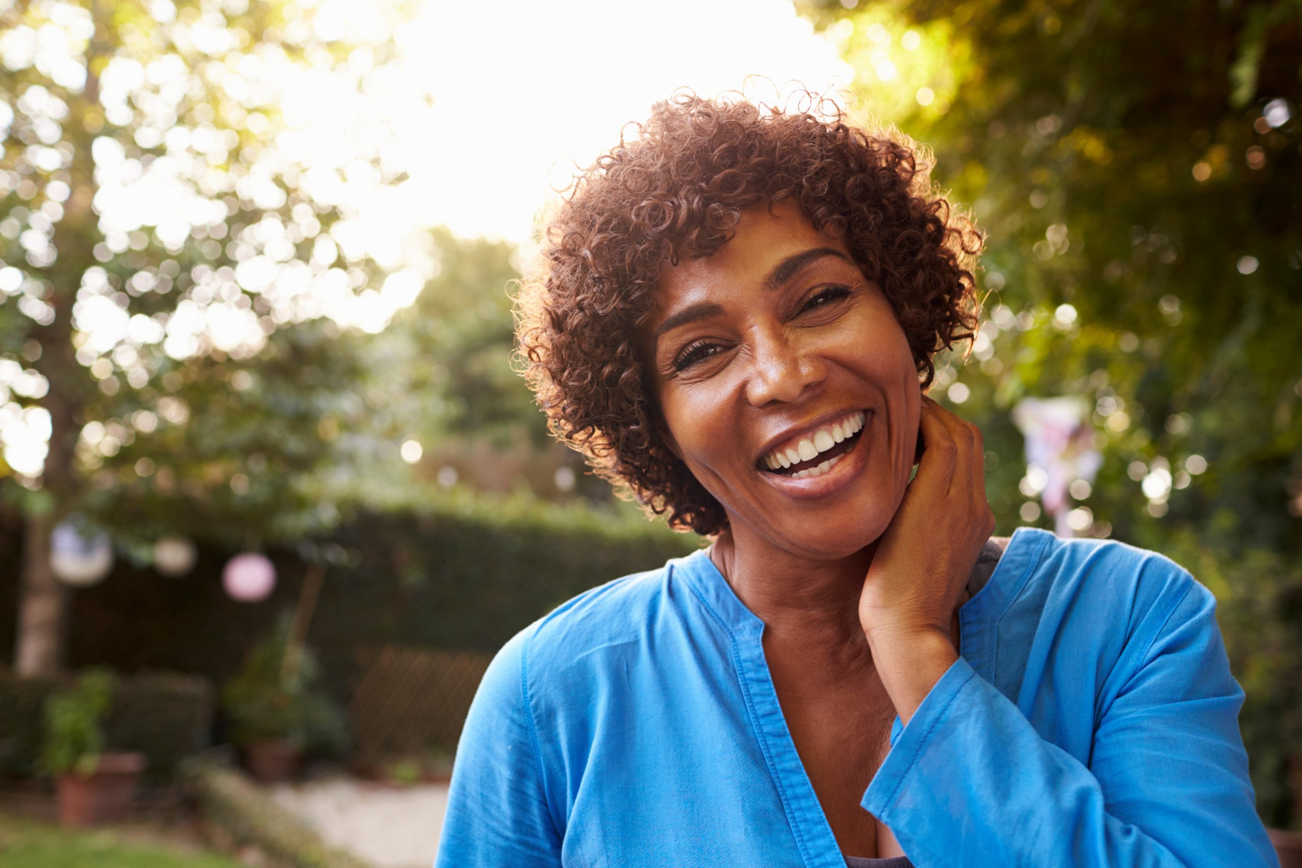 Smiling woman in a blue top standing outdoors with one hand resting on her neck.