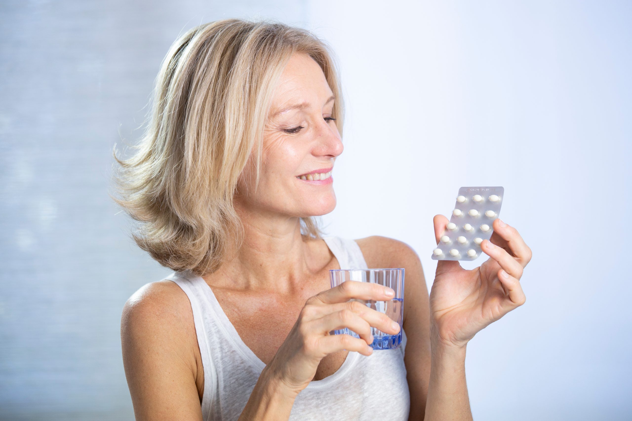 Smiling middle-aged woman holding a glass of water and a blister pack of tablets.