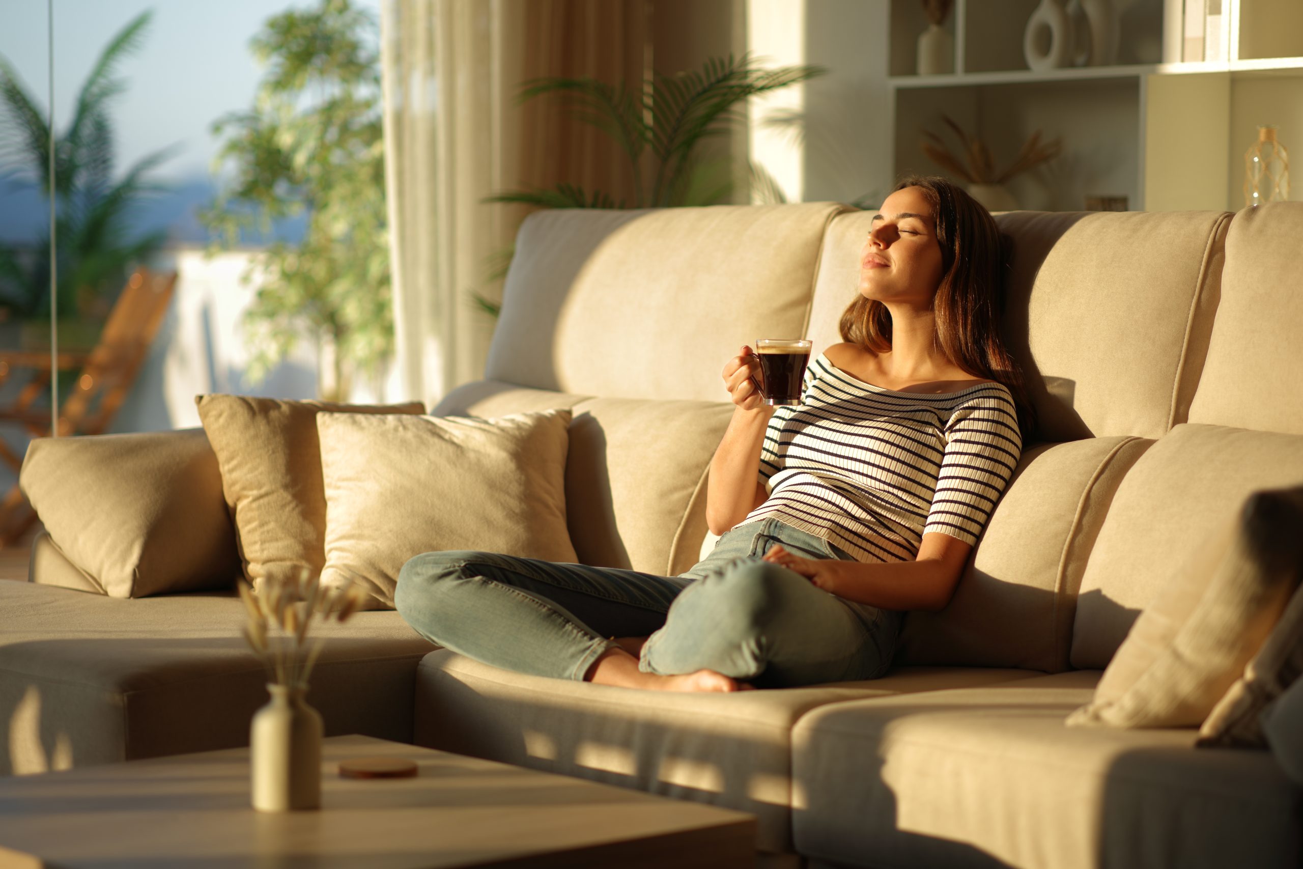 Woman sitting on a couch with her eyes closed, holding a cup of tea and relaxing in a sunlit living room.
